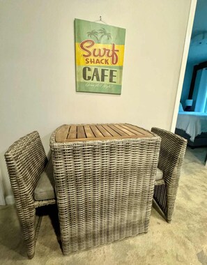 A corner dining area with a wicker table and two chairs, a wall sign reading 'Surf Shack Cafe Open All Night,' and a doorway leading to another room. - Tiny 30A Stay - Walk to Beach Dining Trails (Santa Rosa Beach)
