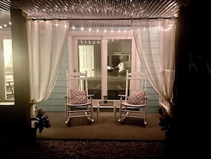 A porch with two rocking chairs, a small table, and a view of a house through a glass door. - Tiny 30A Stay - Walk to Beach Dining Trails (Santa Rosa Beach)