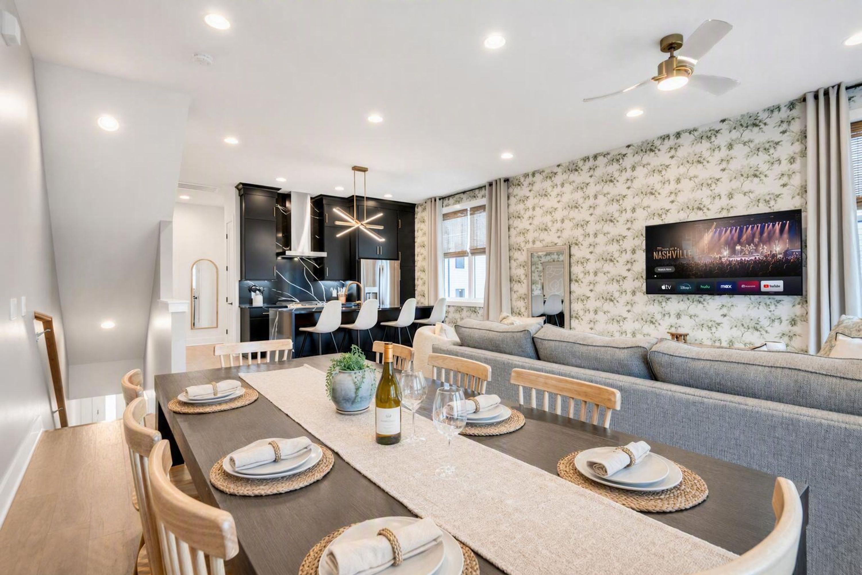A modern living room with a dining area, a flat-screen TV mounted on the wall, and a ceiling fan.