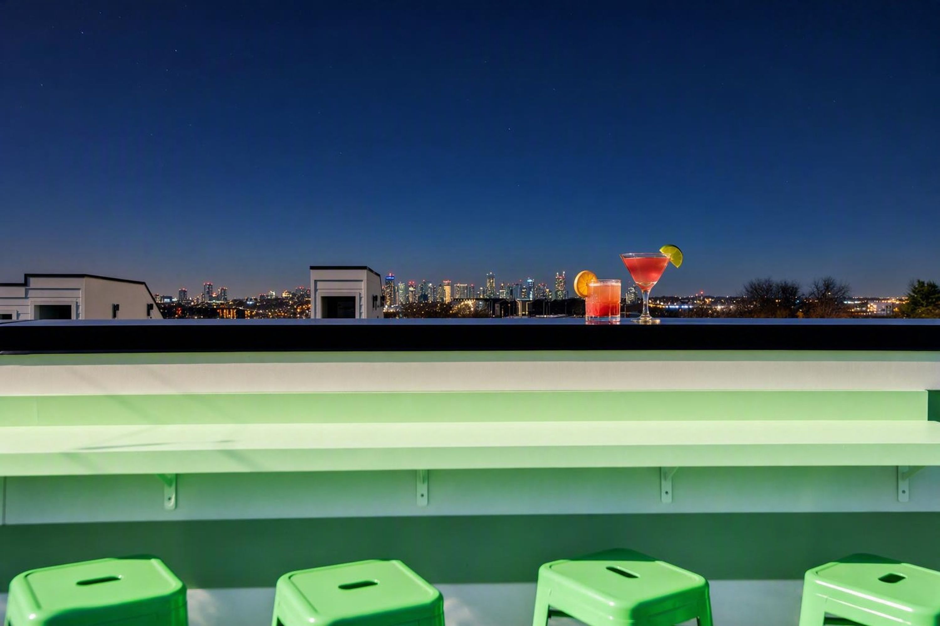 A rooftop bar with a city skyline view, green stools, and two cocktails.