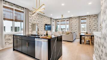A modern kitchen with a central island, a dining area with a wooden table and chairs, and a living area with a sofa and a flat-screen TV.