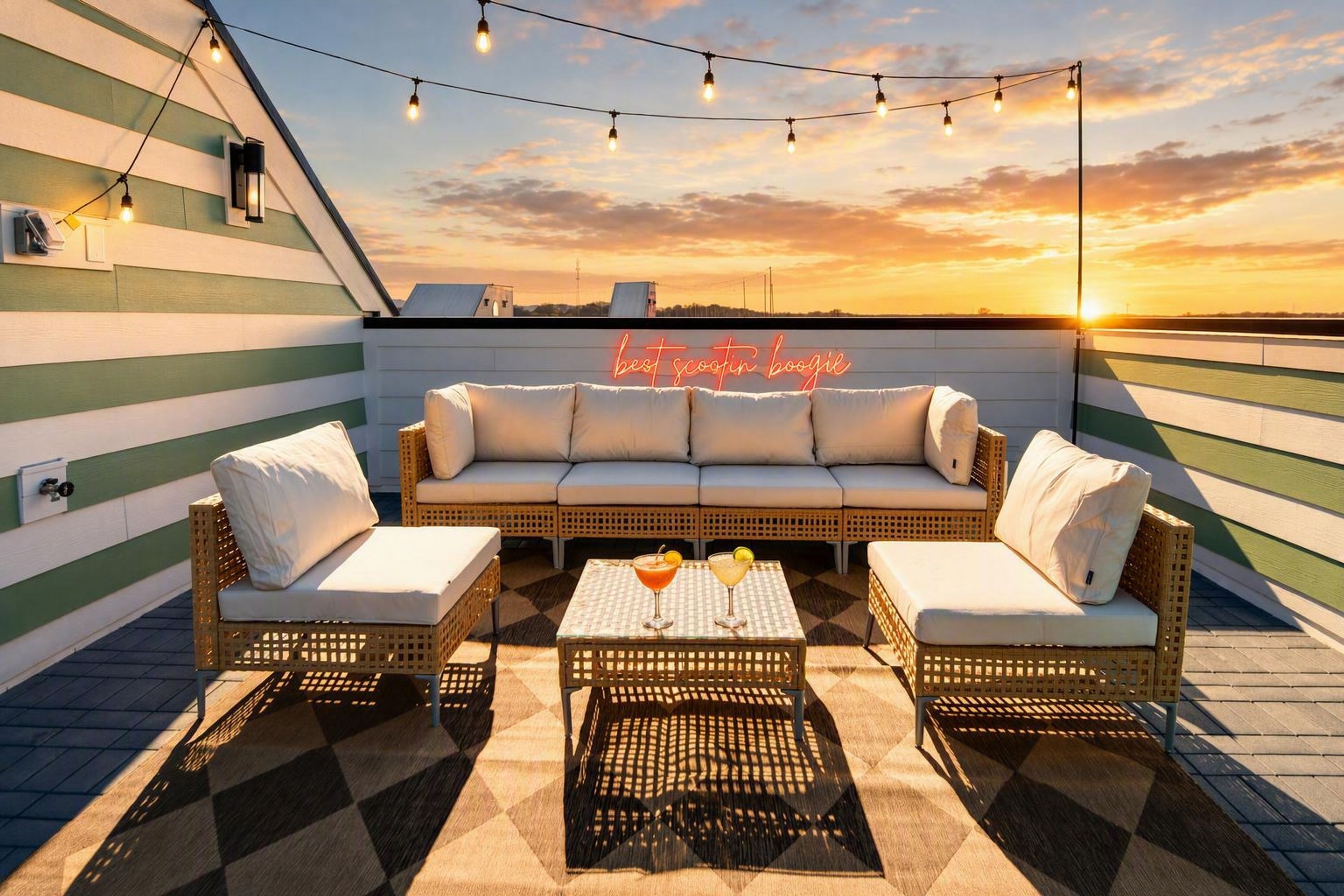 A rooftop terrace with a sofa set, two armchairs, a coffee table, and string lights against a sunset backdrop.