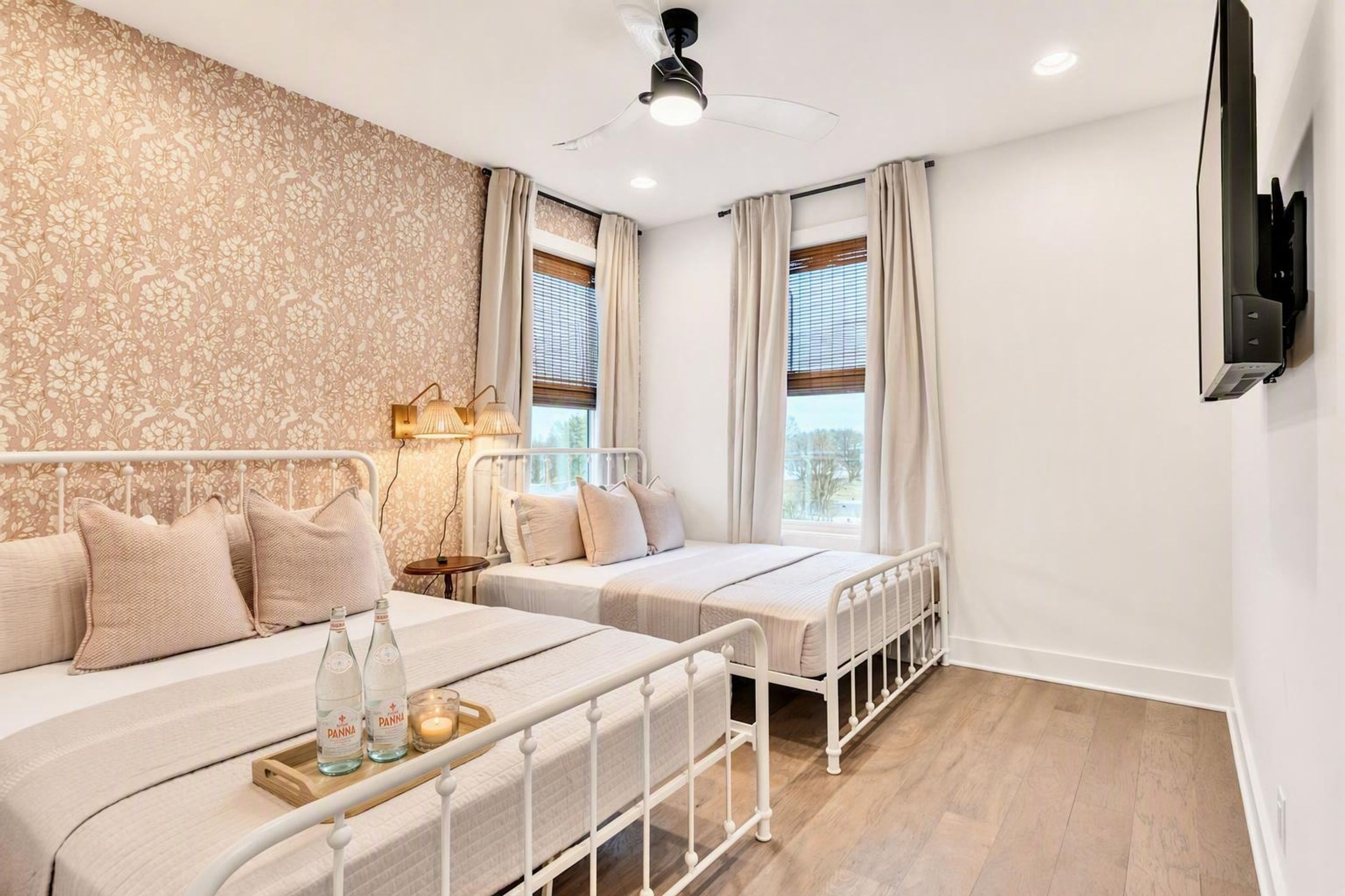 A bedroom with two beds, a ceiling fan, a TV mounted on the wall, and a window with curtains.