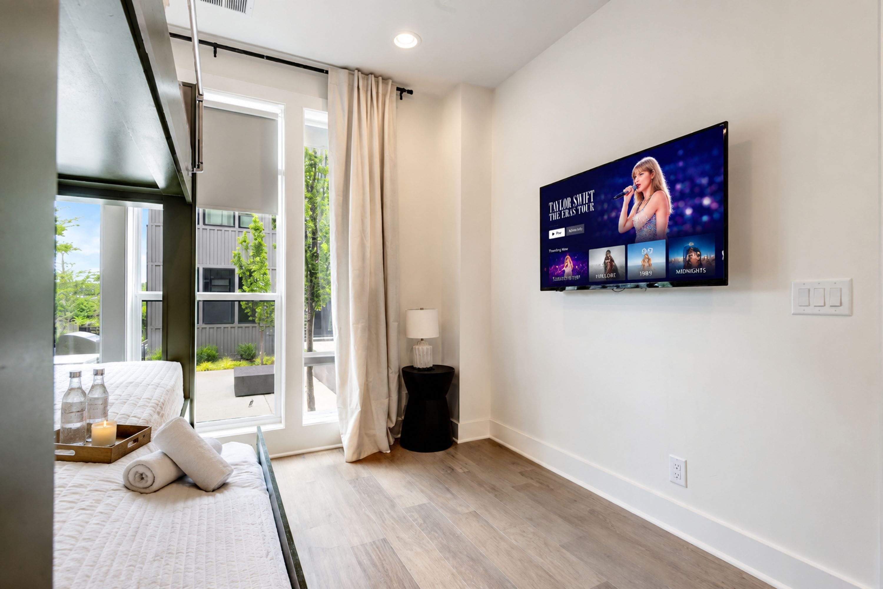A hotel room with a bed, a TV displaying a music artist, a window with curtains, and a view of greenery outside.