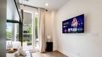 A hotel room with a bed, a TV displaying a music artist, a window with curtains, and a view of greenery outside.