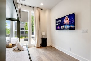 Habitación de hotel con cama, televisor mostrando a un artista musical, ventana con cortinas y vista a espacios verdes al exterior.