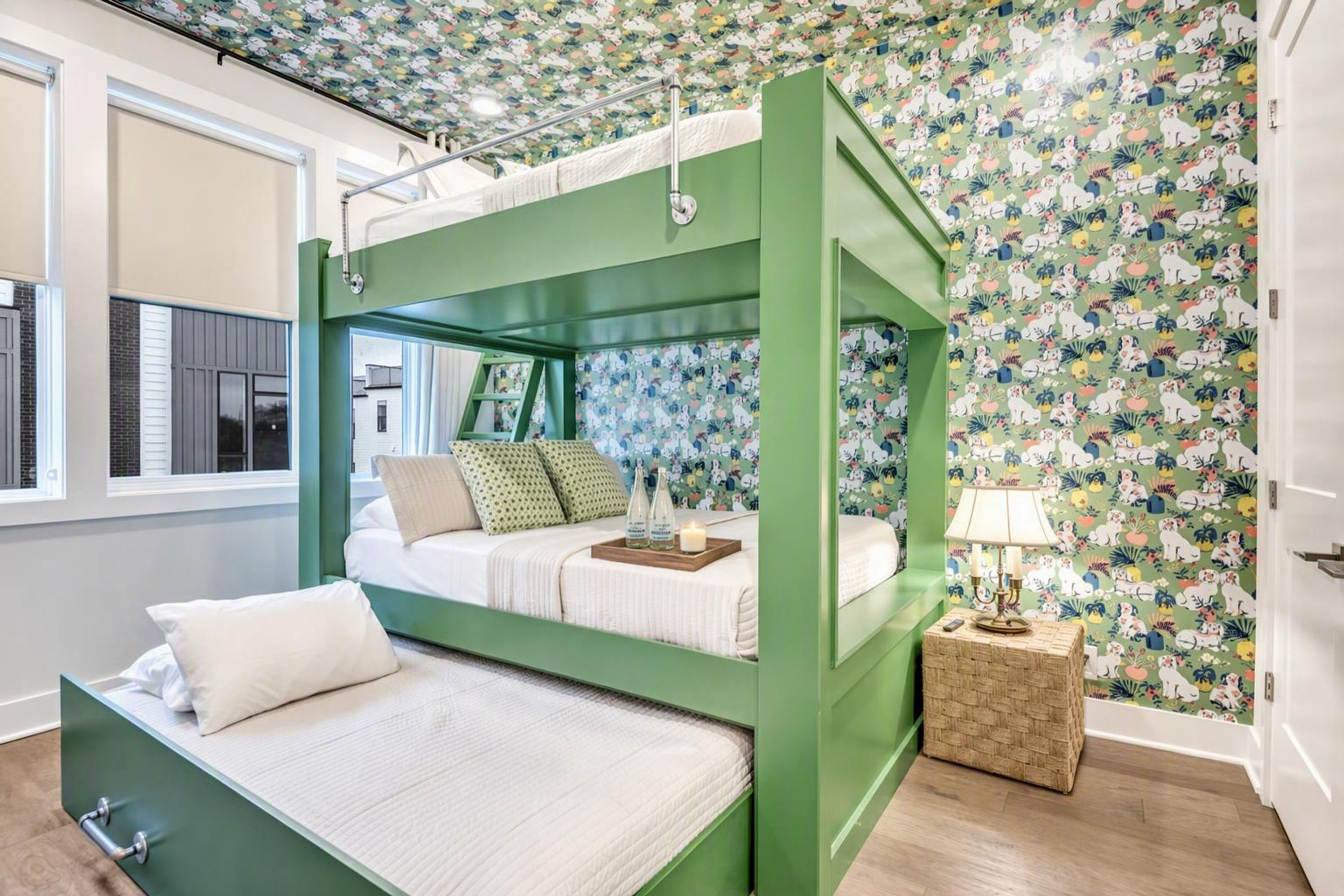 Una cama litera con estructura verde, una mesita de noche con lámpara y una pared con estampado floral.