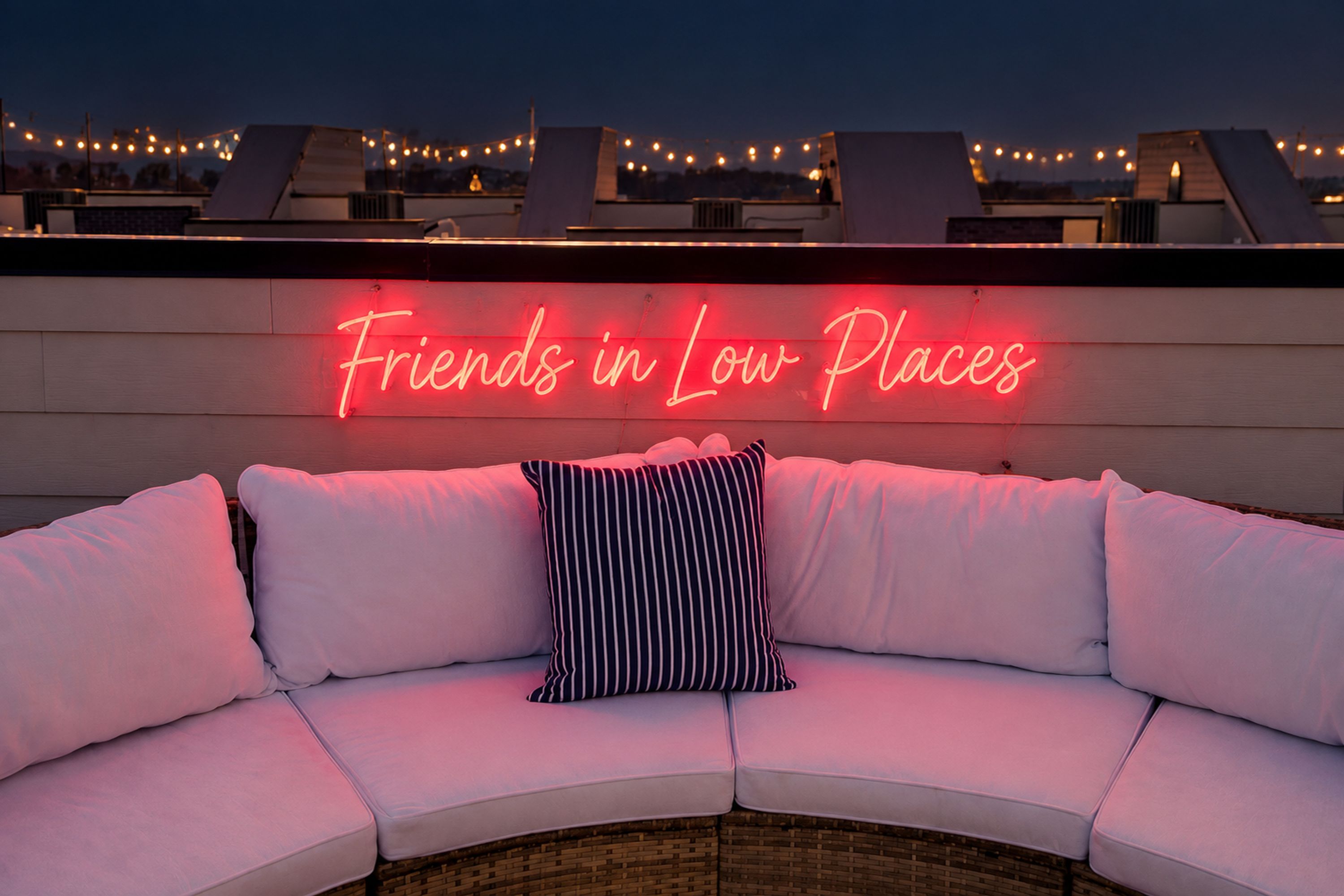 A rooftop seating area with a neon sign reading 'Friends in Low Places' and a white cushioned bench with striped pillows.