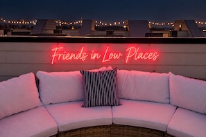 A rooftop seating area with a neon sign reading 'Friends in Low Places' and a white cushioned bench with striped pillows. - Rooftop Deck House Near Geodis Park - Sleeps 12 (Nashville)