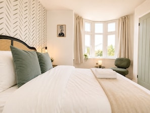 A neatly made bed with pillows, a bedside table with a lamp, a chair, and a window with curtains. - Perfect Booking Modern 4-bed Home With Garden (Windsor)