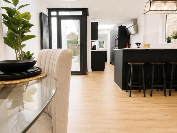 A modern kitchen with a central island, bar stools, and a dining area with a glass table and a chair. - Perfect Booking Modern 4-bed Home With Garden (Windsor)