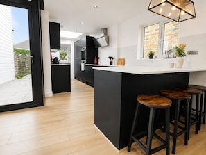 A modern kitchen with a central island, black bar stools, and a window with plants. - Perfect Booking Modern 4-bed Home With Garden (Windsor)
