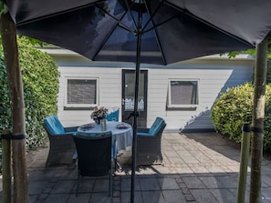 An outdoor patio with a table and chairs under a large umbrella, a white building in the background, and a hedge to the right. - Holiday Home in Renesse With Swimming Pool (Renesse)