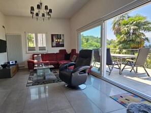 Ein modernes Wohnzimmer mit einem roten Sofa, einem schwarzen Sessel und einem Glastisch. Eine große Schiebetür führt auf eine Terrasse mit Tisch und Stühlen.
