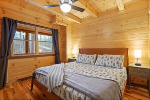 A wooden cabin bedroom with a bed, two bedside lamps, a ceiling fan, and a window with curtains. - Emerald Ridge Log Cabin~Hot Tub~Near Hocking Hills (Logan)