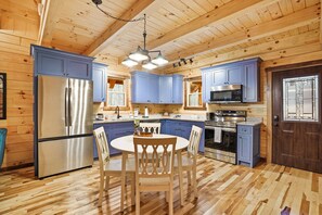 A wooden interior with a kitchen featuring blue cabinets, stainless steel appliances, and a central dining table with chairs. - Sapphire Ridge Log Cabin~Hot Tub~Fire Pit~Hocking (Logan)