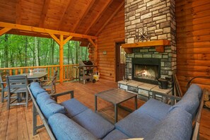 A wooden deck with outdoor seating, a stone fireplace, and a view of the forest. - Sapphire Ridge Log Cabin~Hot Tub~Fire Pit~Hocking (Logan)