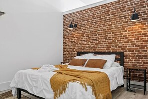 A bedroom with a brick wall, a bed with white bedding and brown pillows, a brown throw blanket, and two black wall-mounted lamps. - Lux Stay Near NYC - Patio, Gym & Free Parking (Kearny)