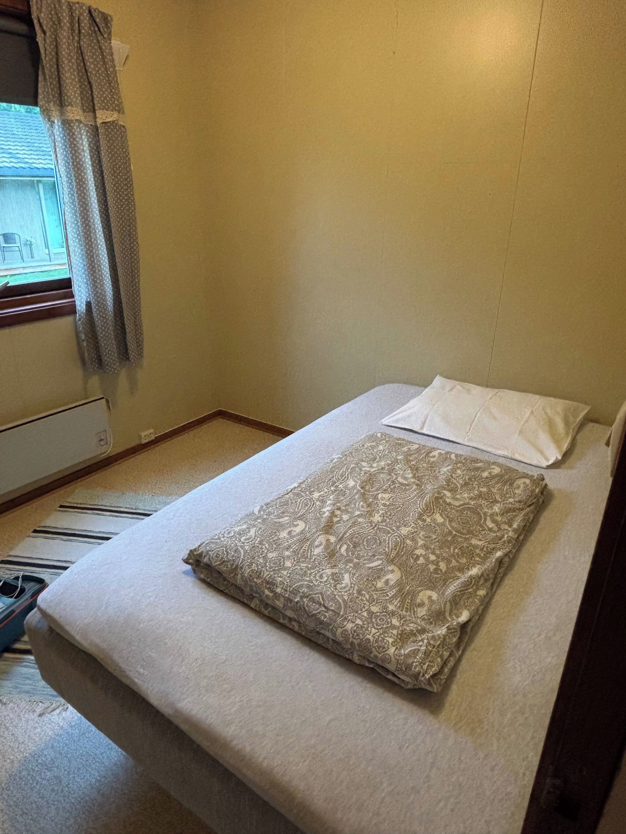 A small, single-bed room with a plain mattress, a pillow, and a window with curtains.