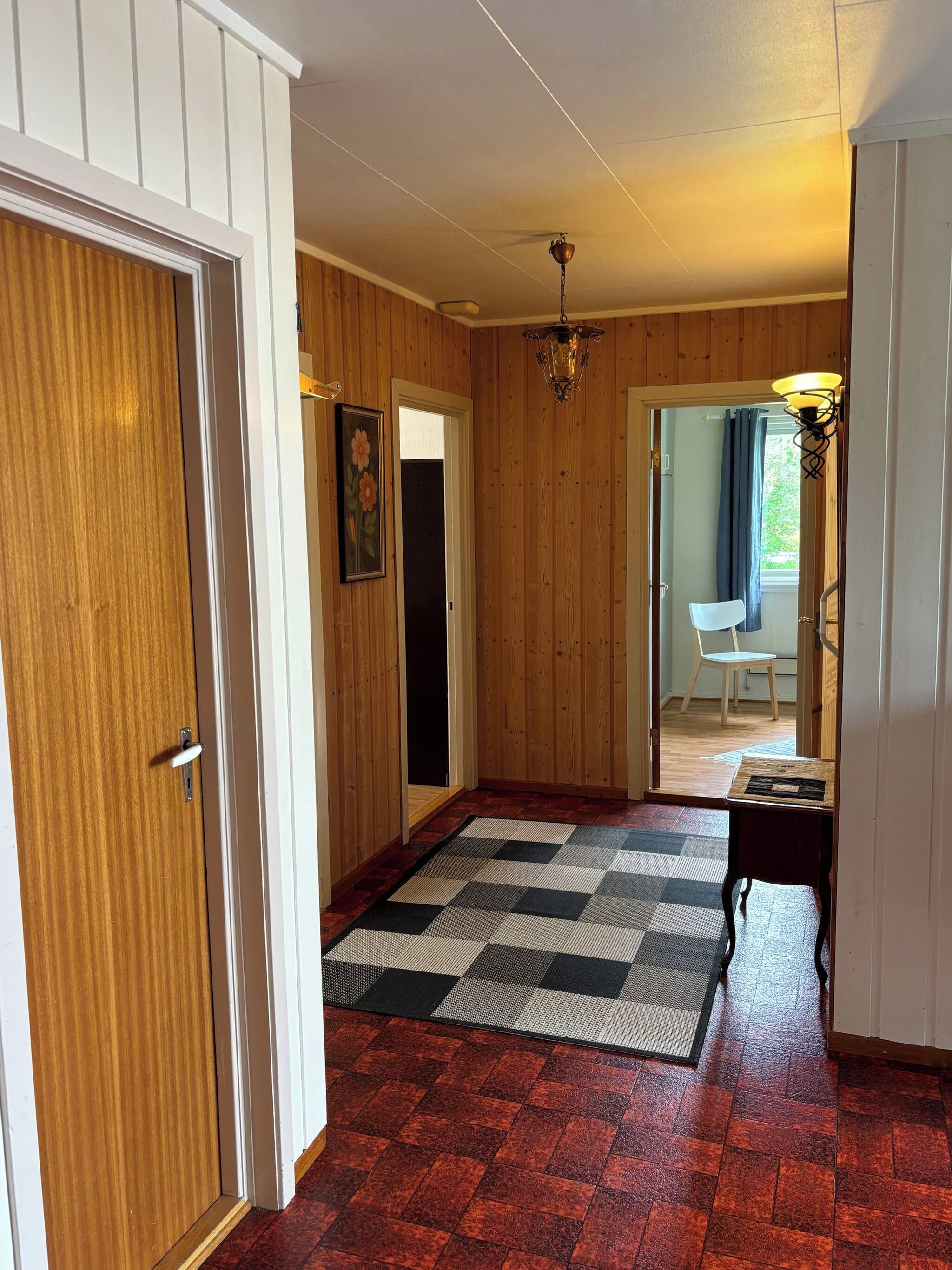 A hallway with wooden walls, a checkered rug, a door with a white handle, and a hanging light fixture.