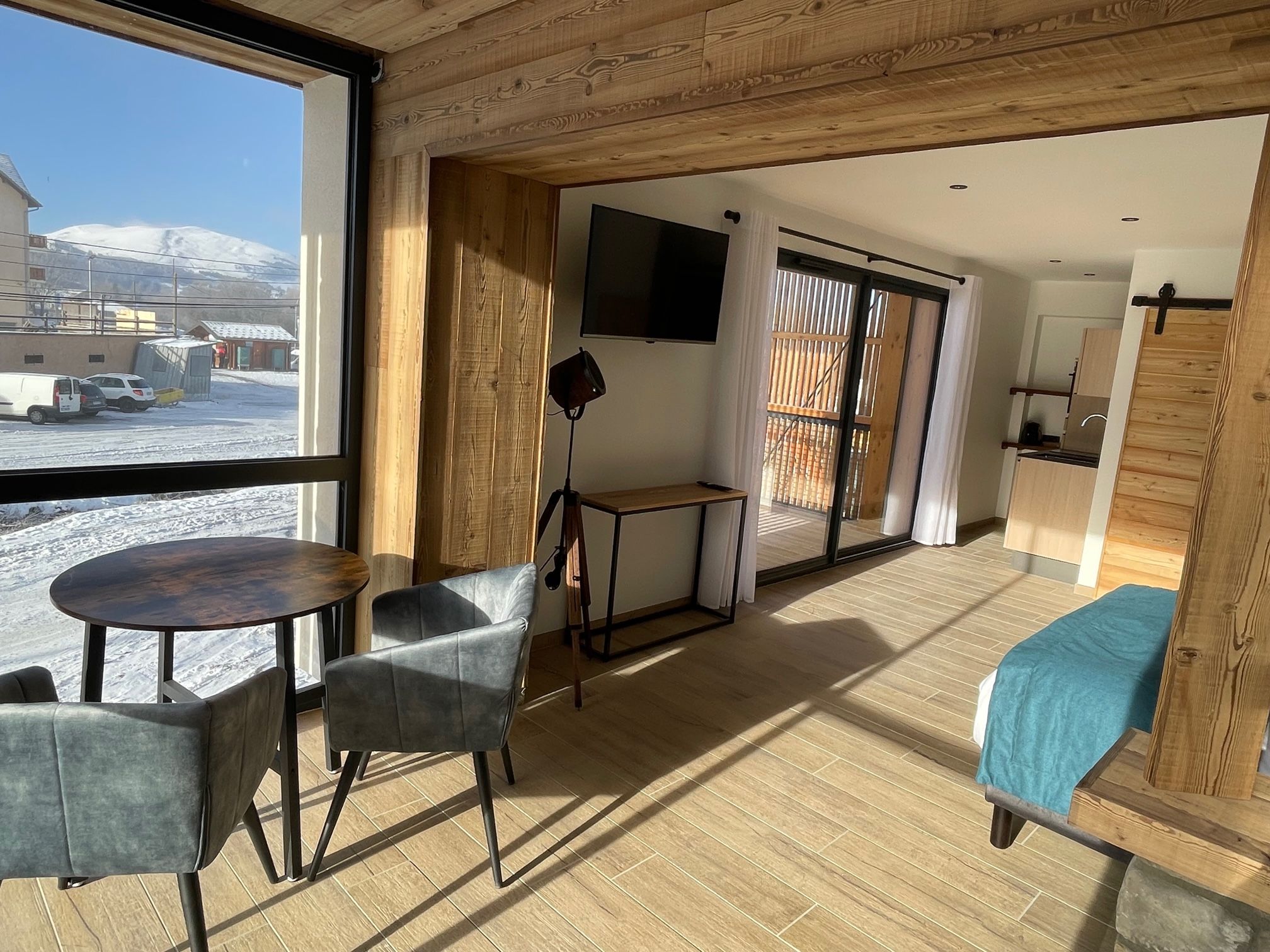 Une pièce avec un plafond en bois, une table ronde entourée de chaises, un lit recouvert d’une couverture bleue, un téléviseur fixé au mur et une vue sur un paysage enneigé par une grande fenêtre.