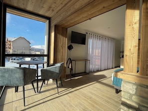 A room with a large window, a wooden ceiling, a TV mounted on the wall, and a seating area with chairs and a table.