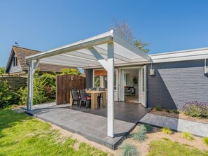A covered patio with a table and chairs, a wooden fence, and a house in the background. - Holiday Home in Renesse With Swimming Pool (Renesse)