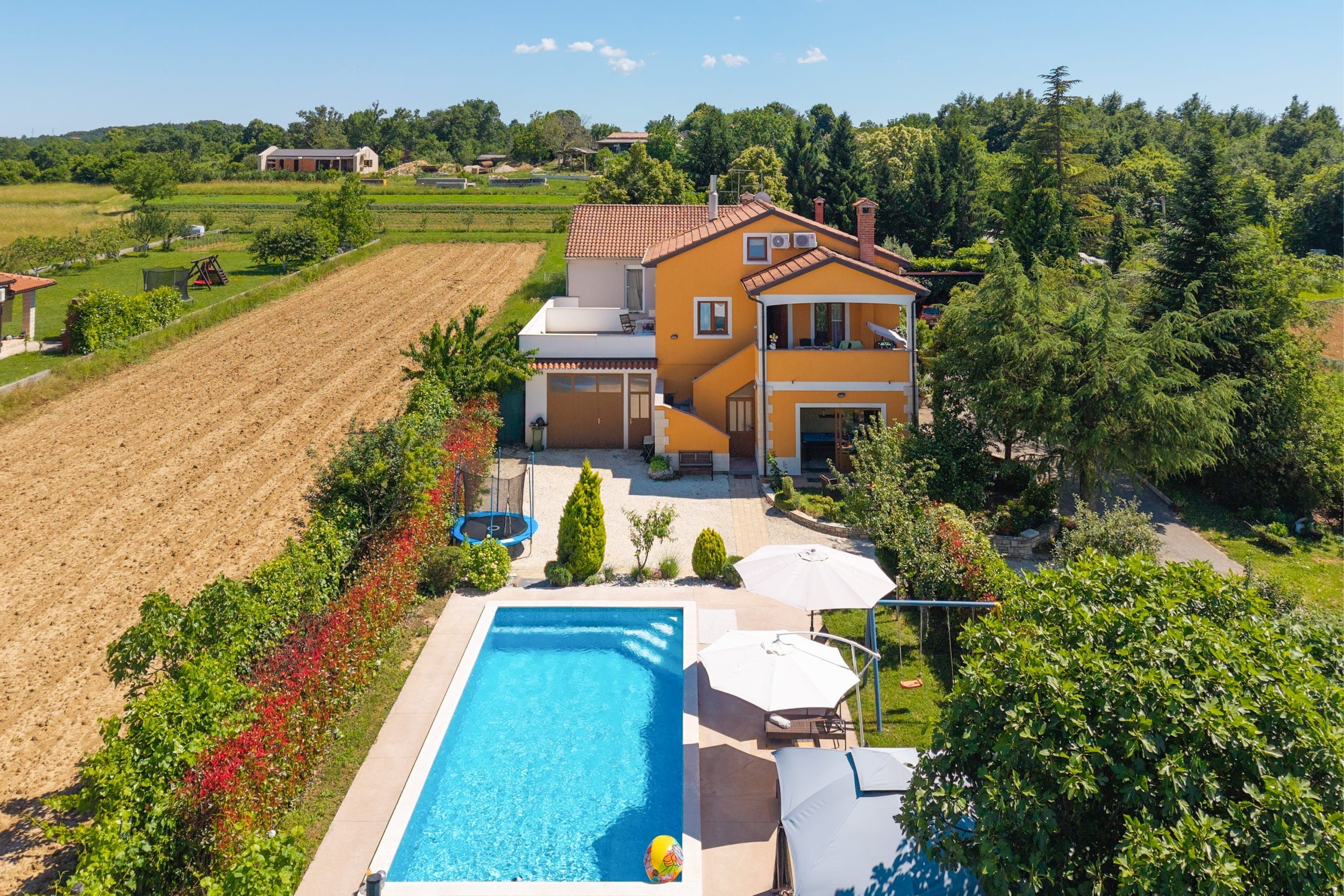 A two-story house with a swimming pool, surrounded by a garden and fields.