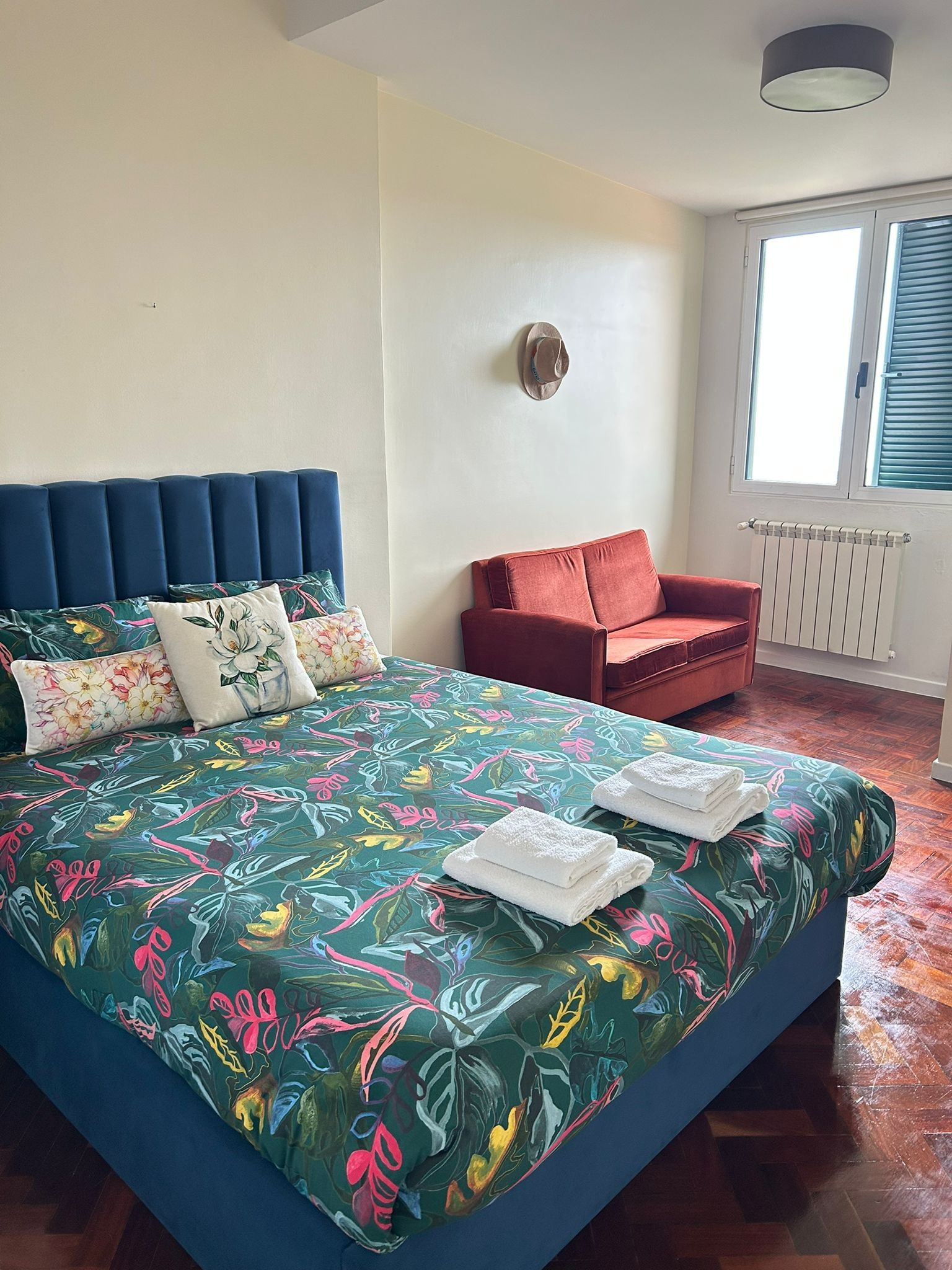 Une chambre à coucher avec un lit, un fauteuil rouge, une fenêtre avec des stores et un radiateur.