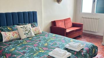 Une chambre à coucher avec un lit, un fauteuil rouge, une fenêtre avec des stores et un radiateur.