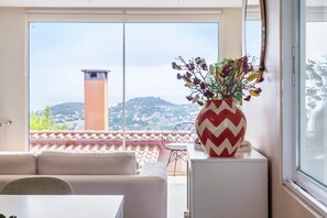 A modern living room with a large window, a white sofa, a dining table, and a decorative vase with artificial flowers.