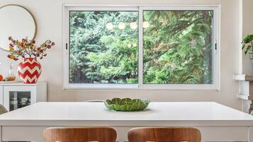 A dining area with a white table, two wooden chairs, a decorative vase with red and yellow flowers, and a window with a view of green foliage.