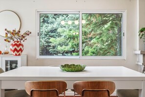 A dining area with a white table, two wooden chairs, a decorative vase with red and yellow flowers, and a window with a view of green foliage.