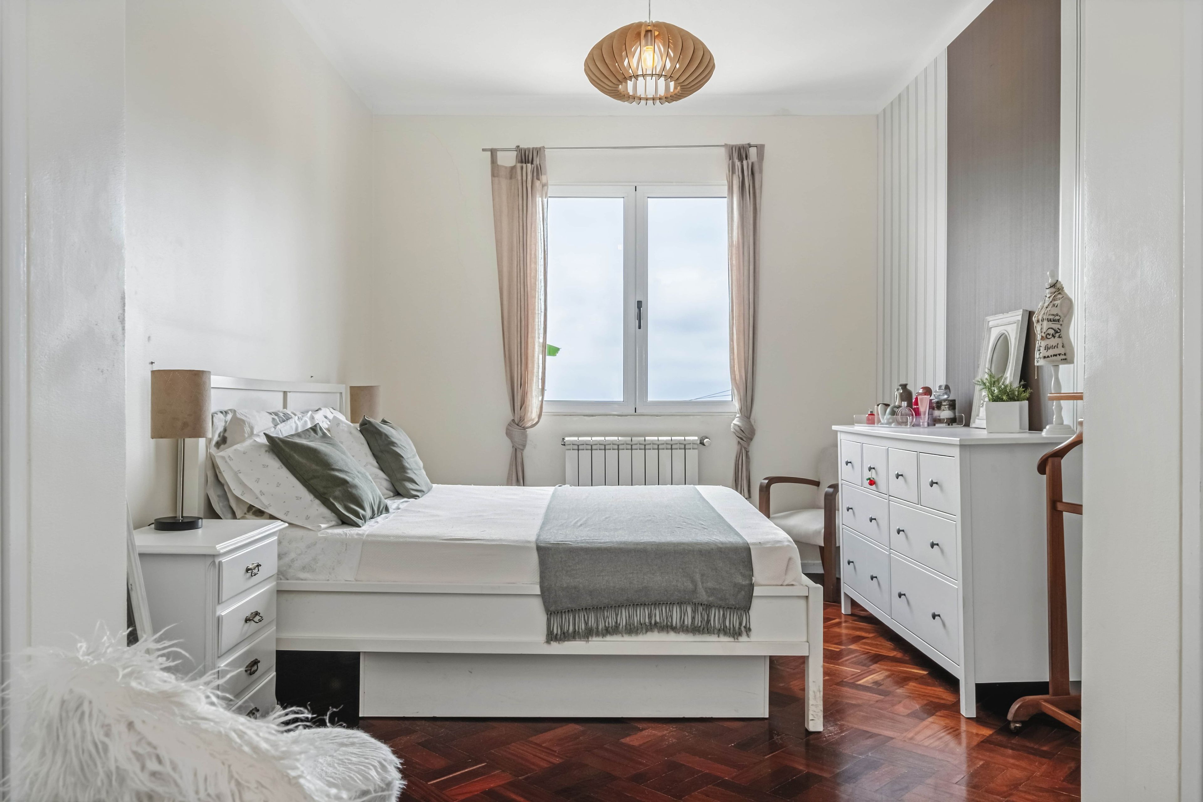 Quarto com cama, mesas de cabeceira, cômoda, radiador e janela com cortinas.