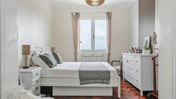 Quarto com cama, mesas de cabeceira, cômoda, radiador e janela com cortinas.