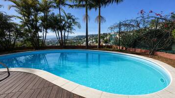 A swimming pool with a wooden deck, surrounded by palm trees and a view of a cityscape.