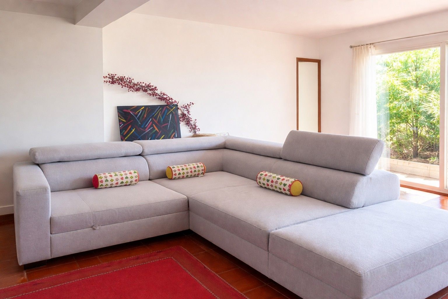 A modern living room with a grey sectional sofa, a red rug, a painting, and a window with a view of greenery.