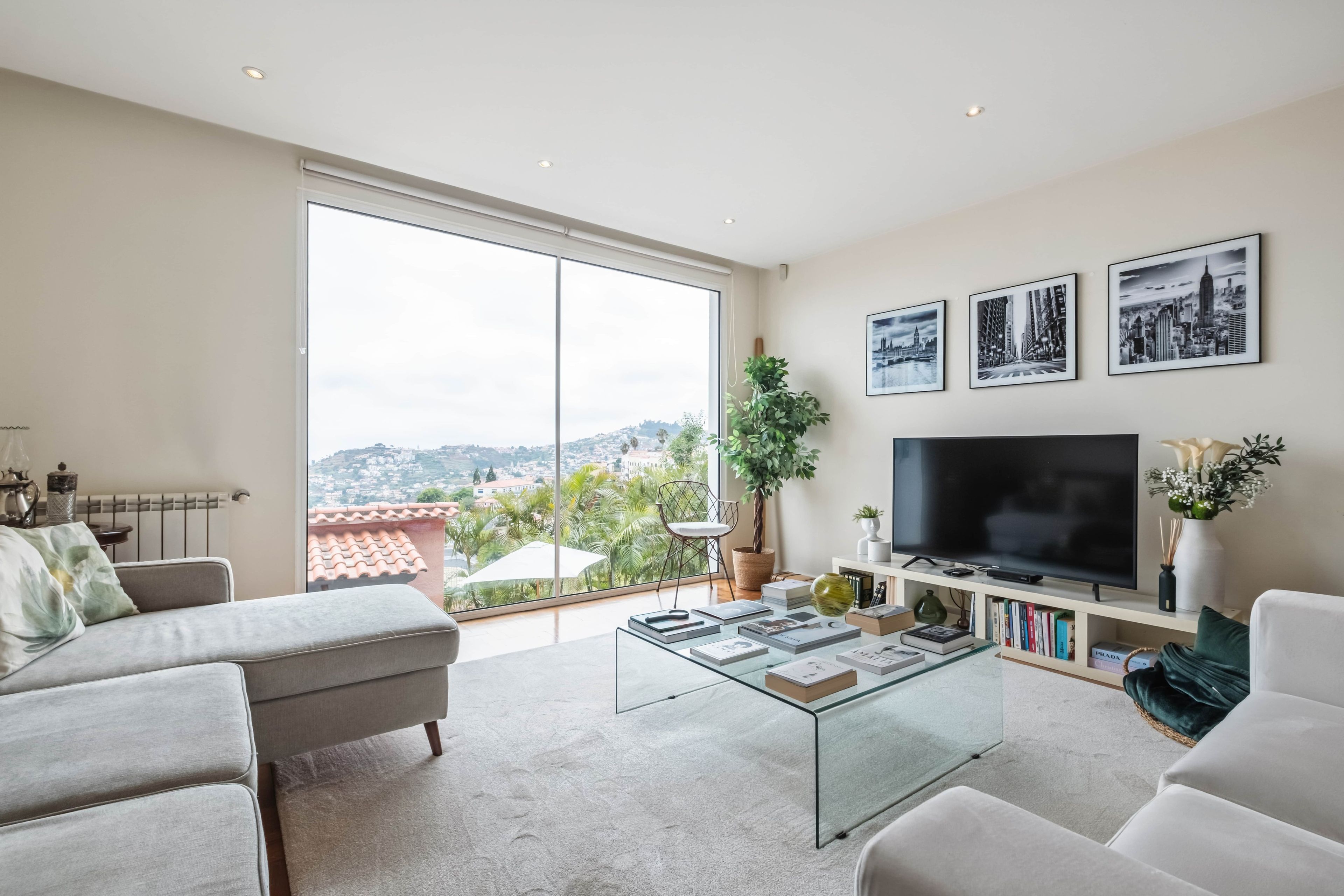 A modern living room with a large window, a flat-screen TV, and a glass coffee table.