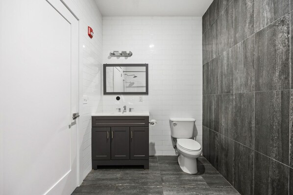 Une salle de bain avec un mur recouvert de carreaux blancs, un meuble-lavabo en bois foncé, des toilettes blanches et un sol carrelé foncé.