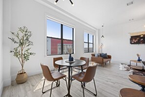 A spacious living area with a round table and chairs, a sofa, and a large window offering a view of a building. - Near NYC High-End Kearny Loft w/ Gym & Patio (Kearny)