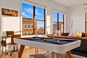 A modern living room with a pool table, a sofa, and a view of the city through large windows. - Expansive 3BR w/ Pool Table - Easy Access to NYC (Kearny)