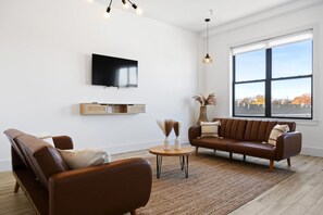 A modern living room with a brown leather sofa, a television mounted on the wall, and a large window providing a view of the outside. - Luxury Stay for Groups Near NYC - Gym & Patio (Kearny)