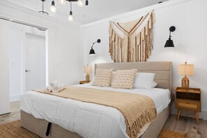 A modern bedroom with a large bed, two bedside tables, a hanging macrame wall decoration, and a door to the left. - Luxury Stay for Groups Near NYC - Gym & Patio (Kearny)