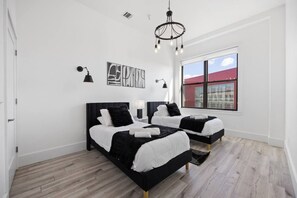 A modern bedroom with two beds, a chandelier, and a large window with a view of a red building. - Huge Top-Floor 2BR, NYC View, Patio & Gym (Kearny)