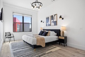 A modern bedroom with a large bed, a side table, a chair, a window with a view, and a chandelier. - Huge Top-Floor 2BR, NYC View, Patio & Gym (Kearny)