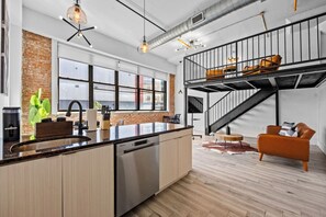 A modern kitchen with a central island, a brick accent wall, and a mezzanine with seating. - Signature Rustic 2BR Near NYC Pets, Gym & Parking (Kearny)