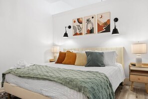 Un dormitorio ordenado con una cama, almohadas, una mesita de noche, una lámpara y decoración en la pared.