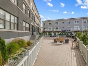 Una terraza de madera con mobiliario exterior, rodeada de edificios con amplias ventanas y un cielo despejado.
