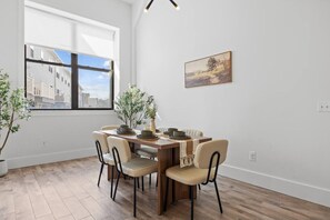 A dining area with a wooden table, six chairs, and a view of buildings through a large window. - 20 Mins to NYC Chic 2BR Loft w/ Gym & BBQ (Kearny)