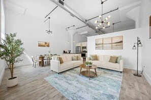 A modern living room with a sofa, coffee table, and a dining area with chairs and a table. - 20 Mins to NYC Chic 2BR Loft w/ Gym & BBQ (Kearny)